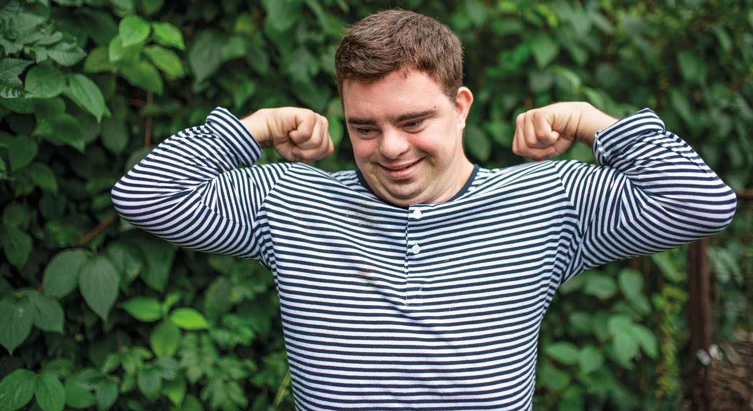 Ein erwachsener Mann mit Down-Syndrom, der stolz seine Muskeln beugt, vor einem grünen Hintergrund im Freien. Down-Syndrom